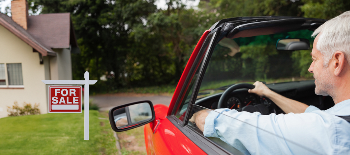 A man in a red convertible driving by a home with a 'For Sale' out in front.