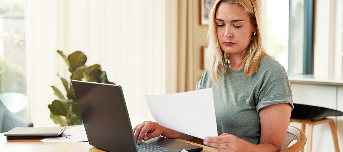 A woman sitting at a table holding up a piece of paper in front of an open laptop.