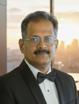 A formal portrait of a man with a mustache and glasses, wearing a black tuxedo with a black bowtie. He is looking directly at the camera with a neutral expression. The background shows a large window with a blurred view of a city skyline during sunset, casting a warm, golden light over the scene.