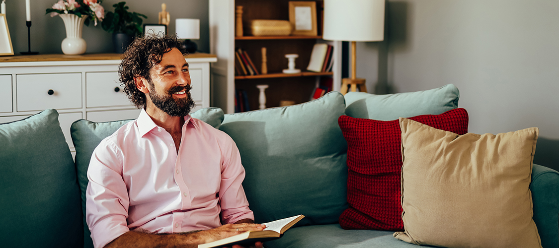 A man sits smiling and reading a book on his couch