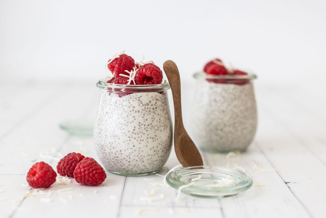 Two chia puddings in glass jars are featured on a white wooden surface. The chia puddings are topped with fresh raspberries and sprinkled with shredded coconut.