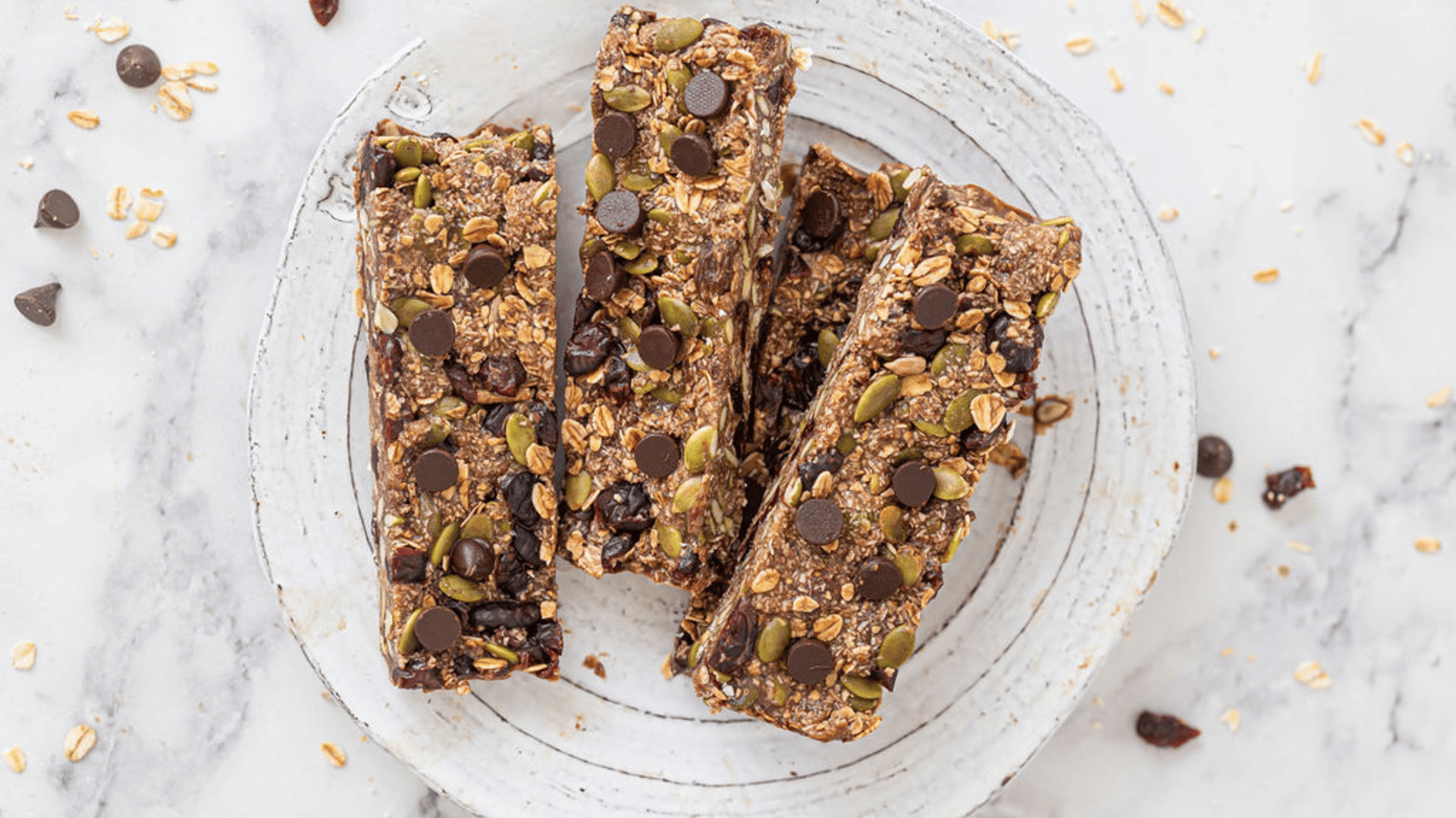 Three homemade granola bars are placed upon baking paper. The bars are sprinkled with pepitas, dark chocolate drops and shredded coconut.