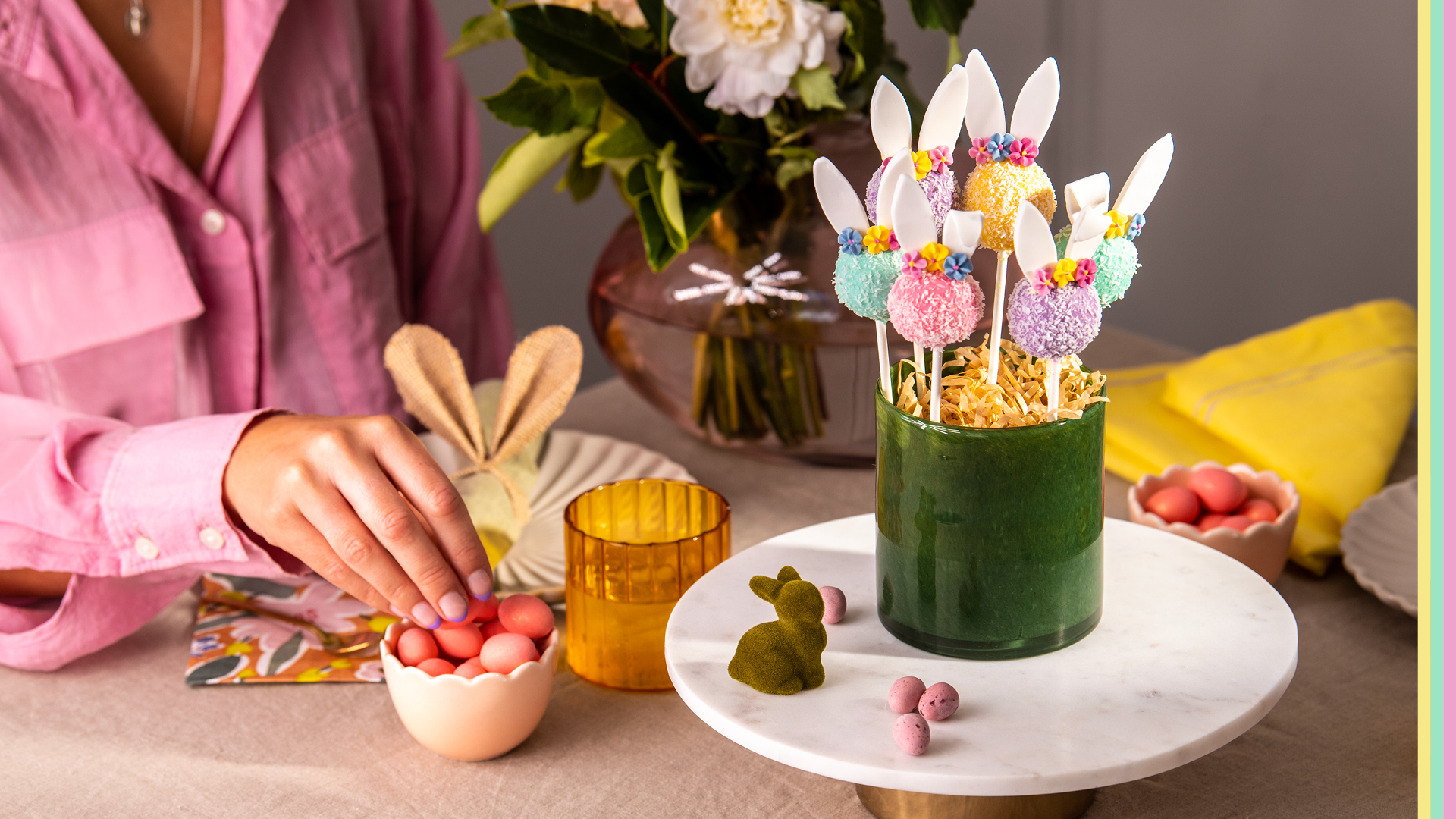 Multi coloured Easter bunny cake pops rolled in desiccated coconut displayed in dark green vase with sugar Easter eggs in background