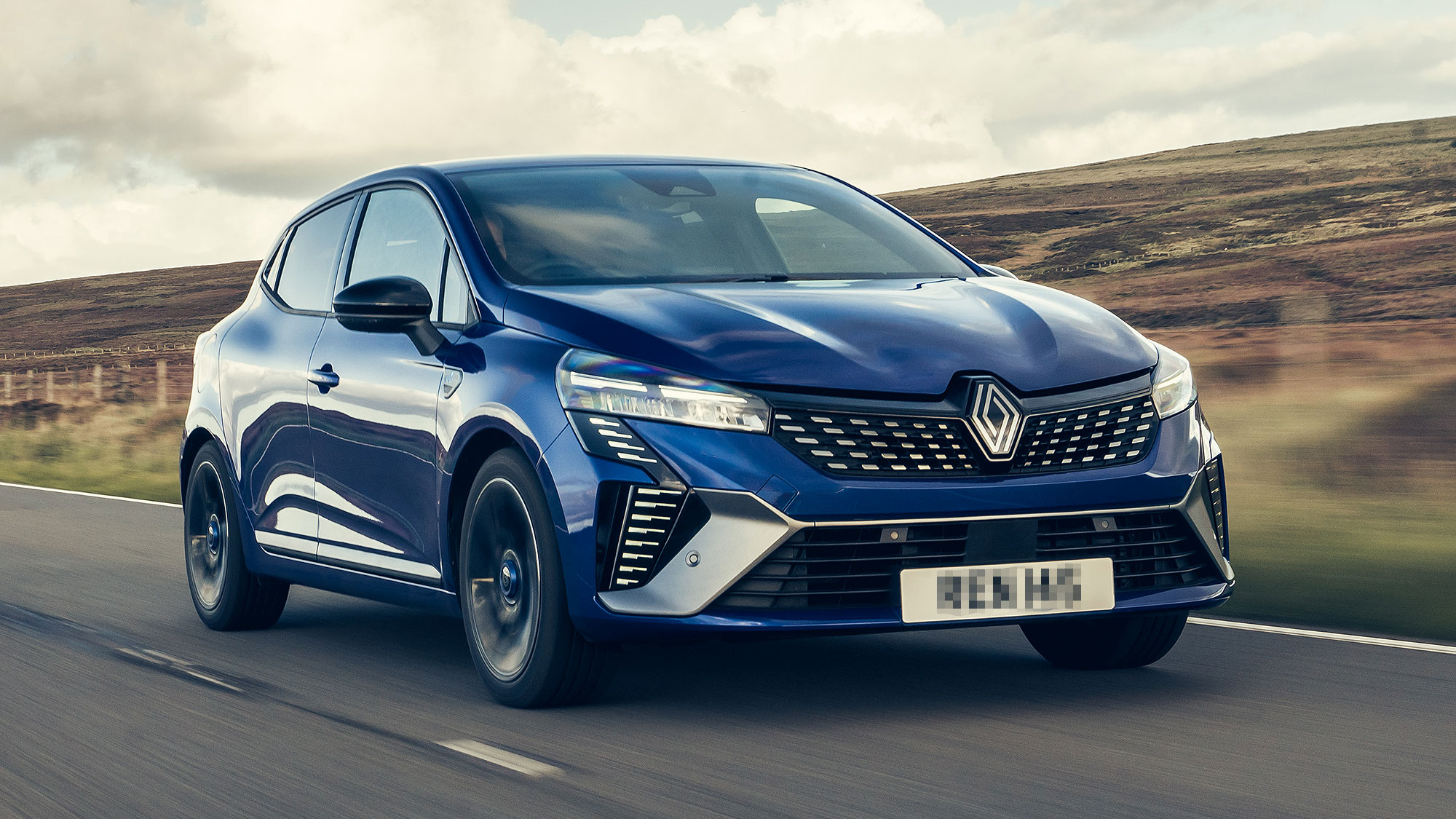 A metallic blue Renault Clio driving along a rural road, showcasing slim LED headlights and a sculpted front design.
