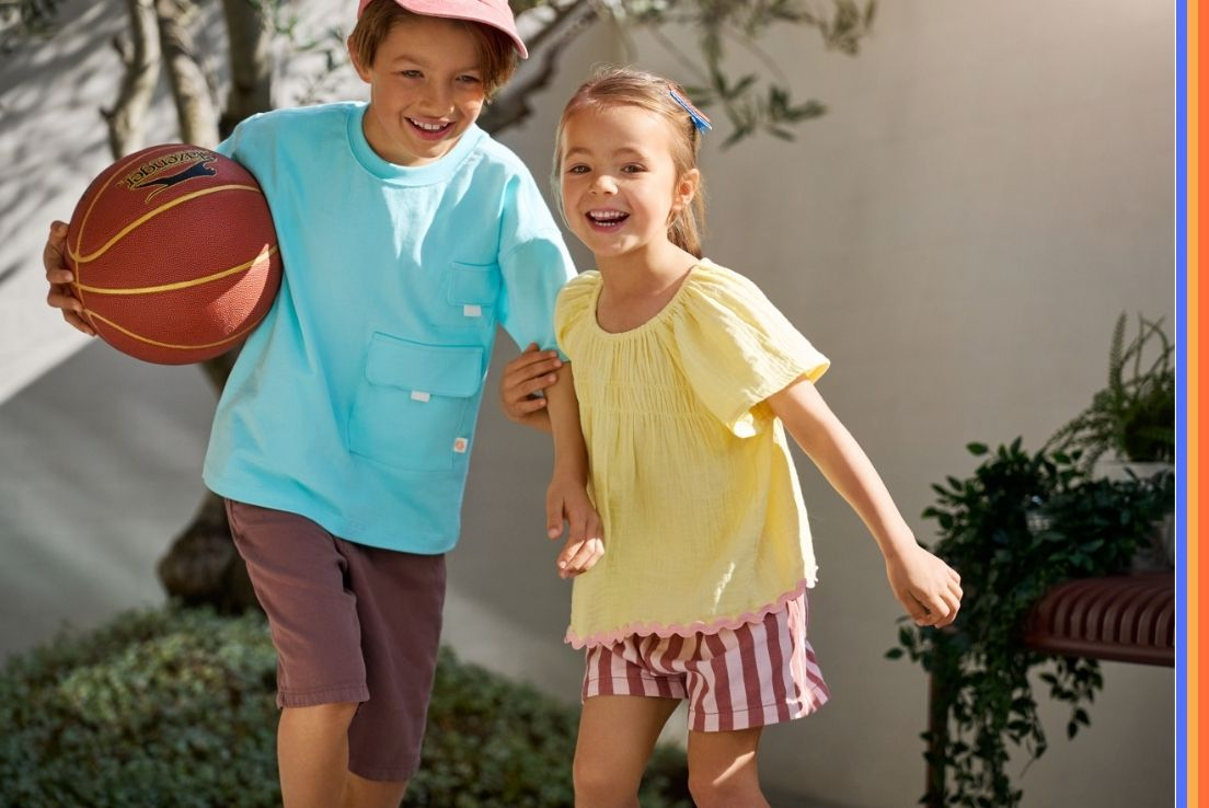 Two children play outdoors - the boy wears a pink cap and holds a basketball; the girl wears a yellow top and striped shorts.