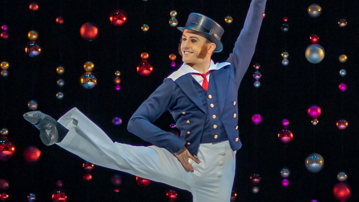 A dancer dressed in a blue and white sailor-style costume performs a high kick on stage with colourful baubles hanging in the background.