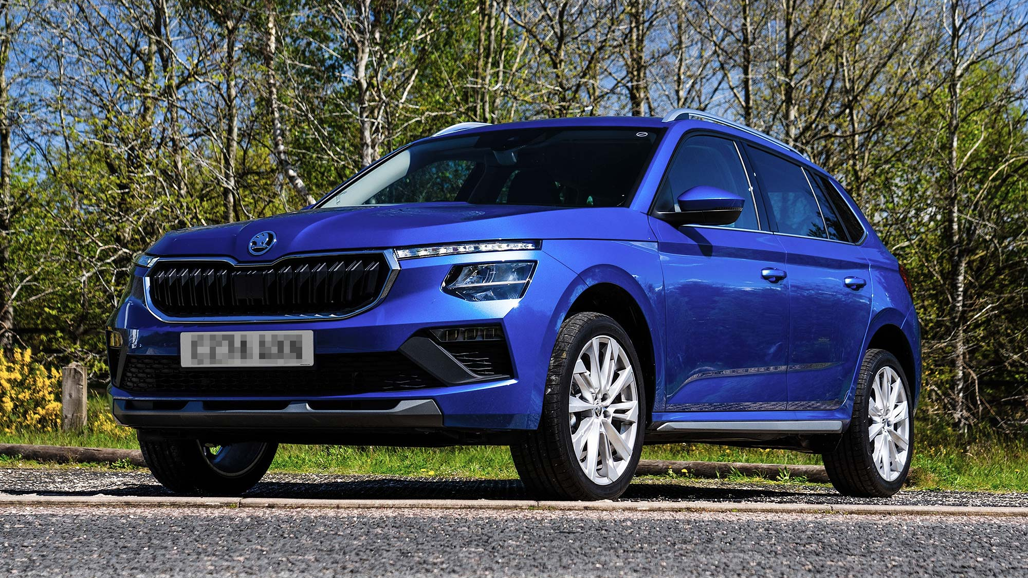 Blue Škoda Kamiq compact SUV parked on roadside tarmac, with spring trees and greenery in the background.
