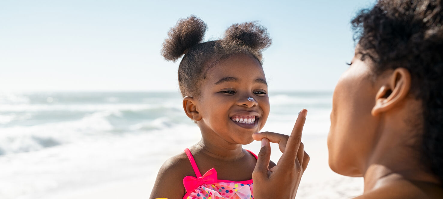 Young African American mother applying protective sunscreen on daughter's nose at beach.