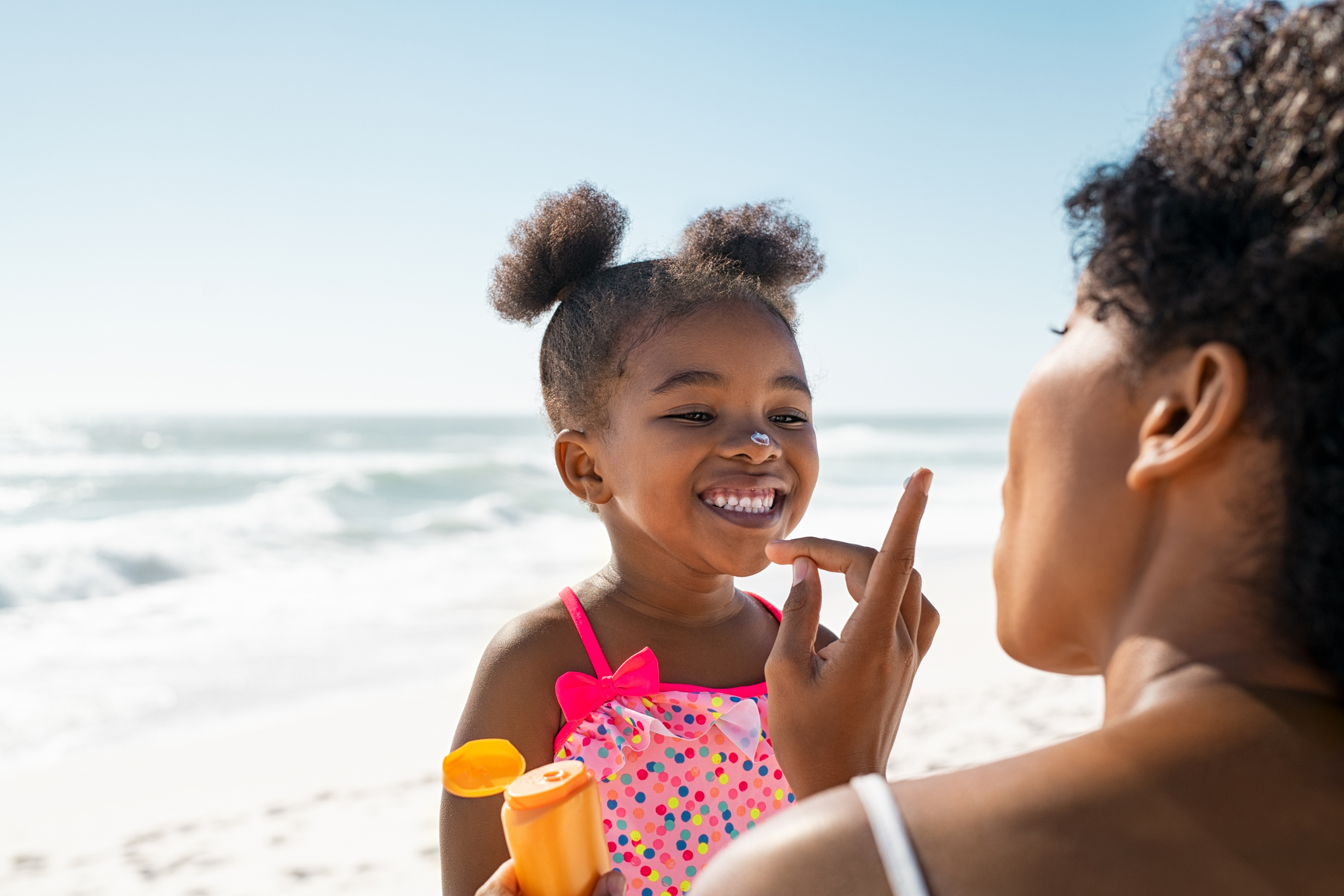 Young African American mother applying protective sunscreen on daughter's nose at beach.