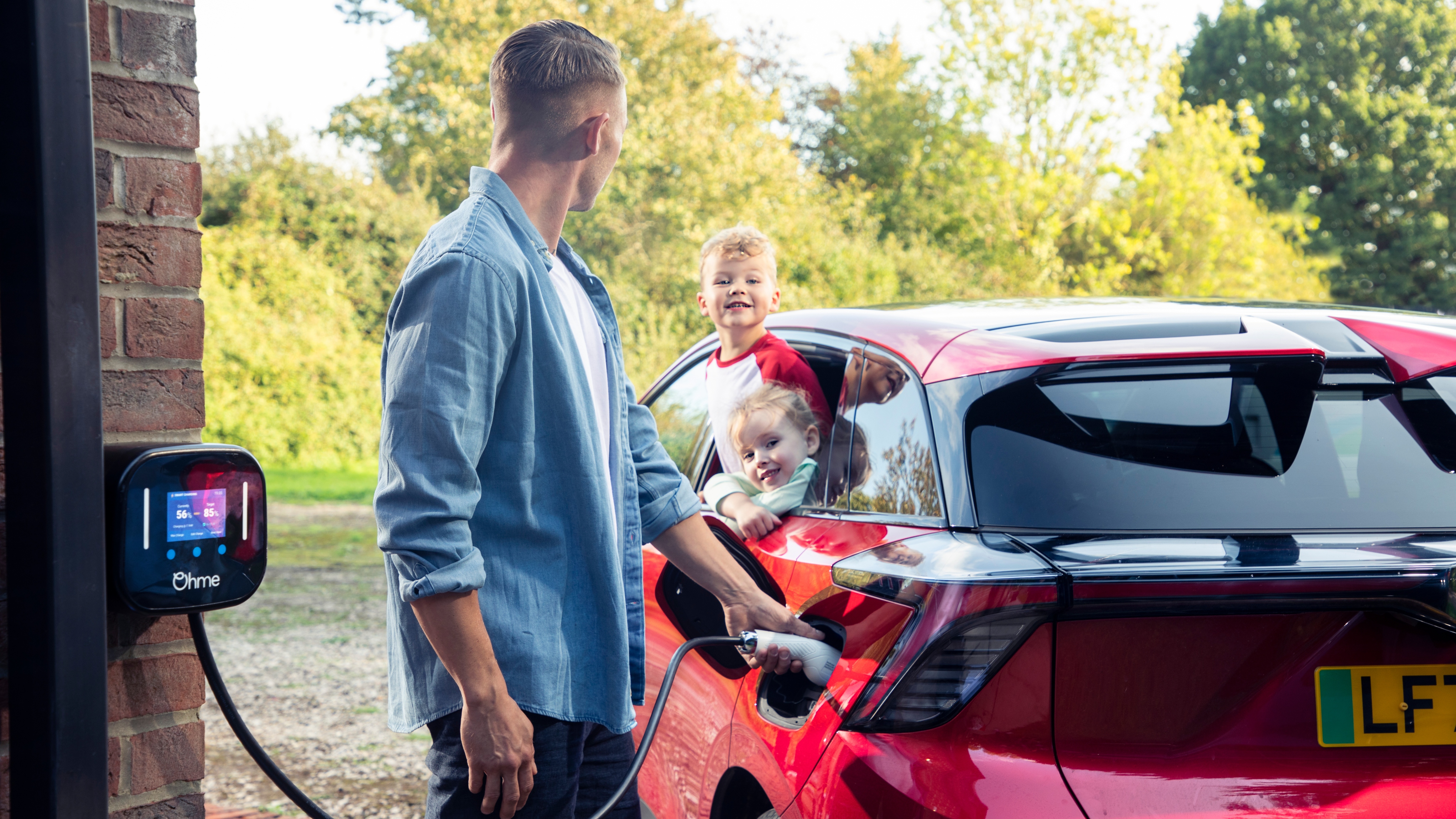 A man plugs a red electric car into a home charging point while two children smile and lean out of the rear window, with trees and greenery in the background.
