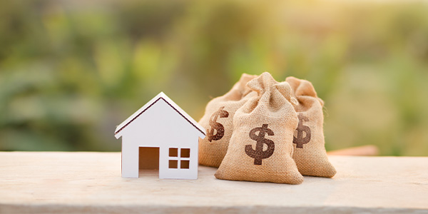 A small wooden model of a house and two little bags with a money symbol on them sit on a desk top 