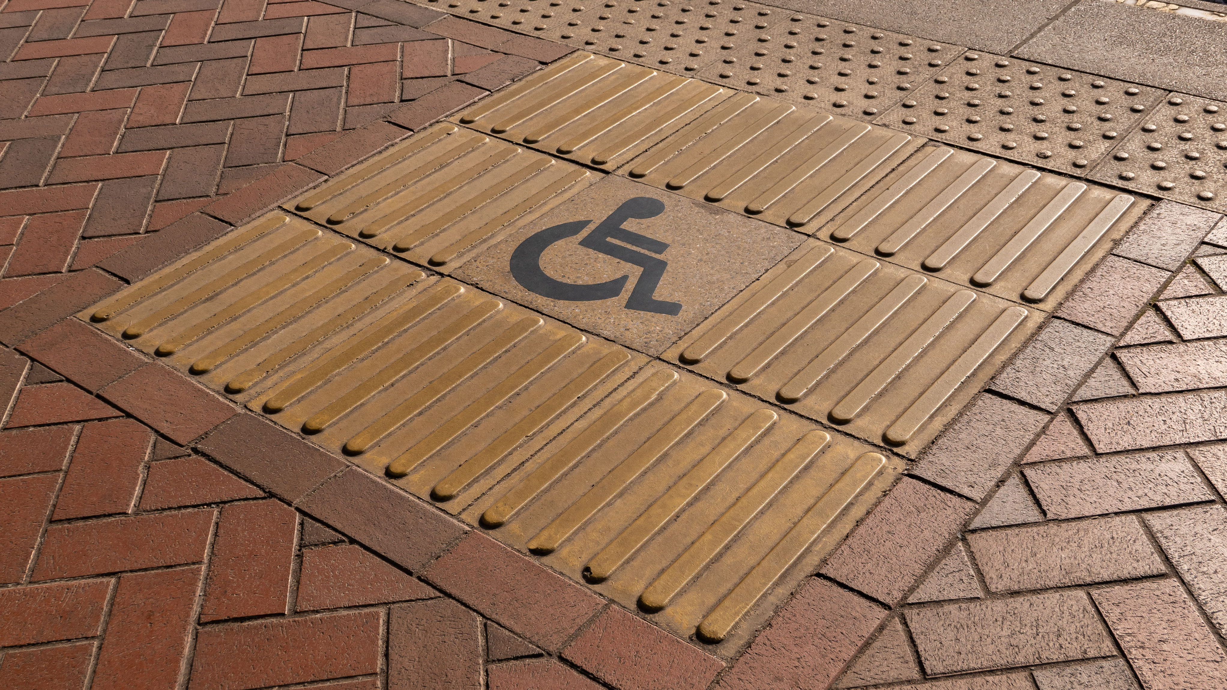A section of pavement with tactile paving featuring raised ridges and a wheelchair accessibility symbol in the center, designed to assist visually impaired and disabled pedestrians.