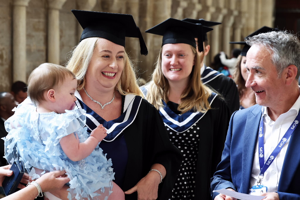 Two female graduates in caps and gowns, one holding a baby in a light blue frilly outfit