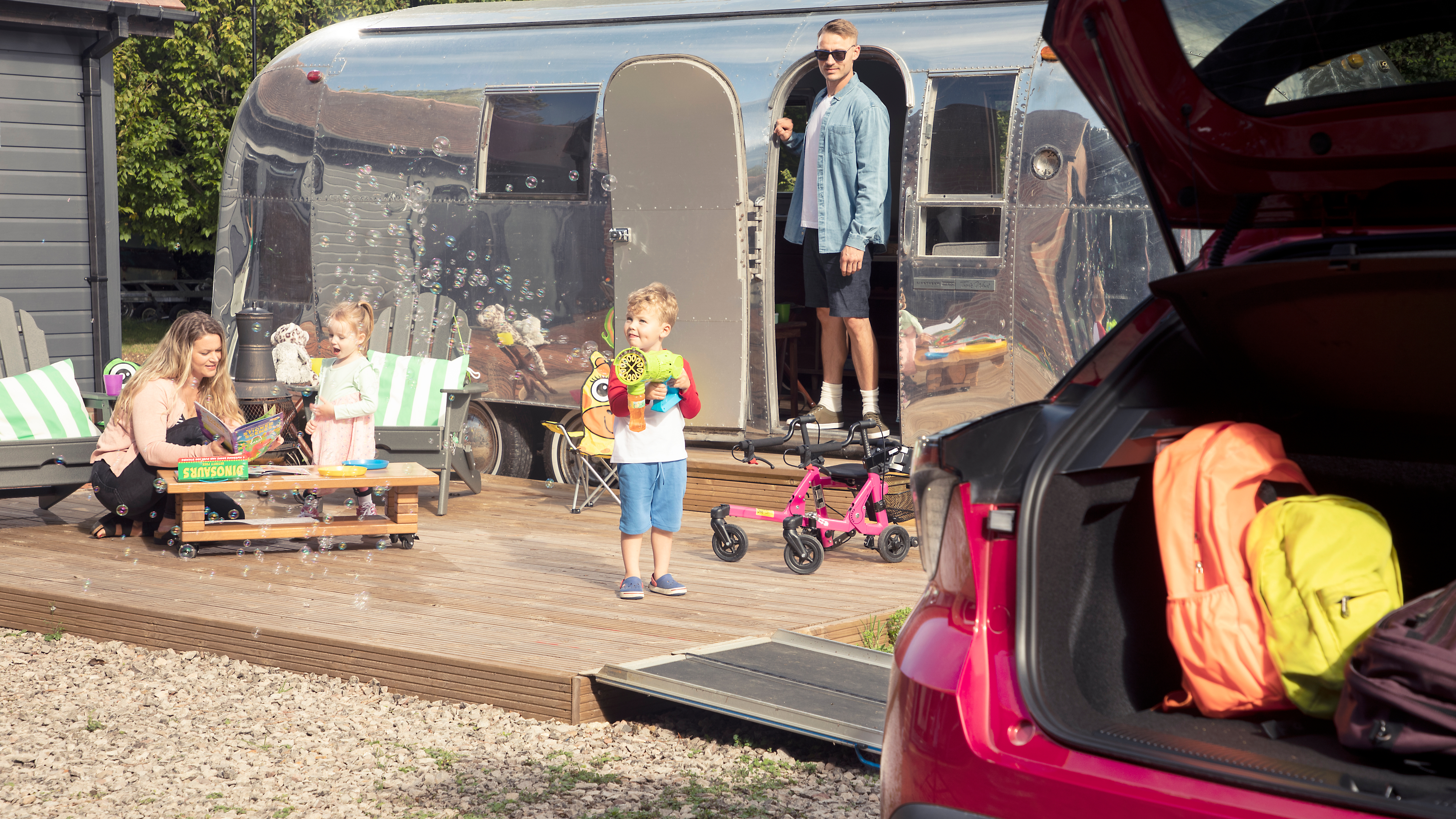 A family enjoying time outside a silver caravan on a sunny day. A young boy stands on a wooden deck blowing bubbles with a toy, while a little girl plays nearby. A woman sits at a low table reading with children’s books and toys around her. A man stands in the open doorway of the caravan, looking on. In the foreground, the boot of a red car is open, showing colourful backpacks packed inside, suggesting they’ve just arrived or are setting off on a trip.