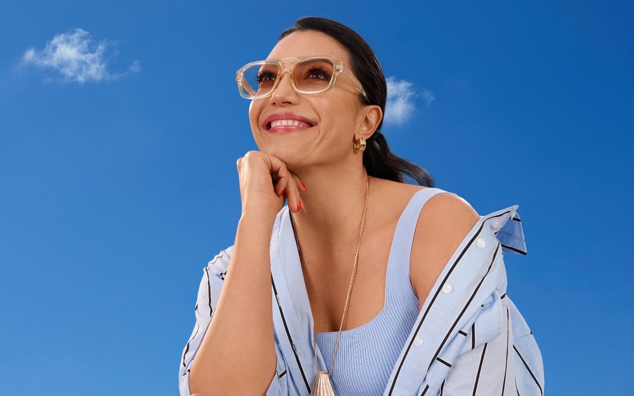 Smiling woman wearing sunglasses and a light blue top with a striped jacket draped over her shoulders, posing against a bright blue sky.