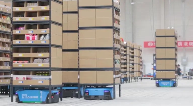 Automated guided vehicles (AGVs) carrying tall stacks of cardboard boxes and metal shelving units in a brightly lit, expansive warehouse. The warehouse floor is clear, and the AGVs, which are low-profile and blue/teal, navigate between rows of shelving. On the left, some shelves hold various products, while the tall units on the AGVs are mostly stacked with large, uniform brown cardboard boxes. In the background, there are more stacks and a few human workers.