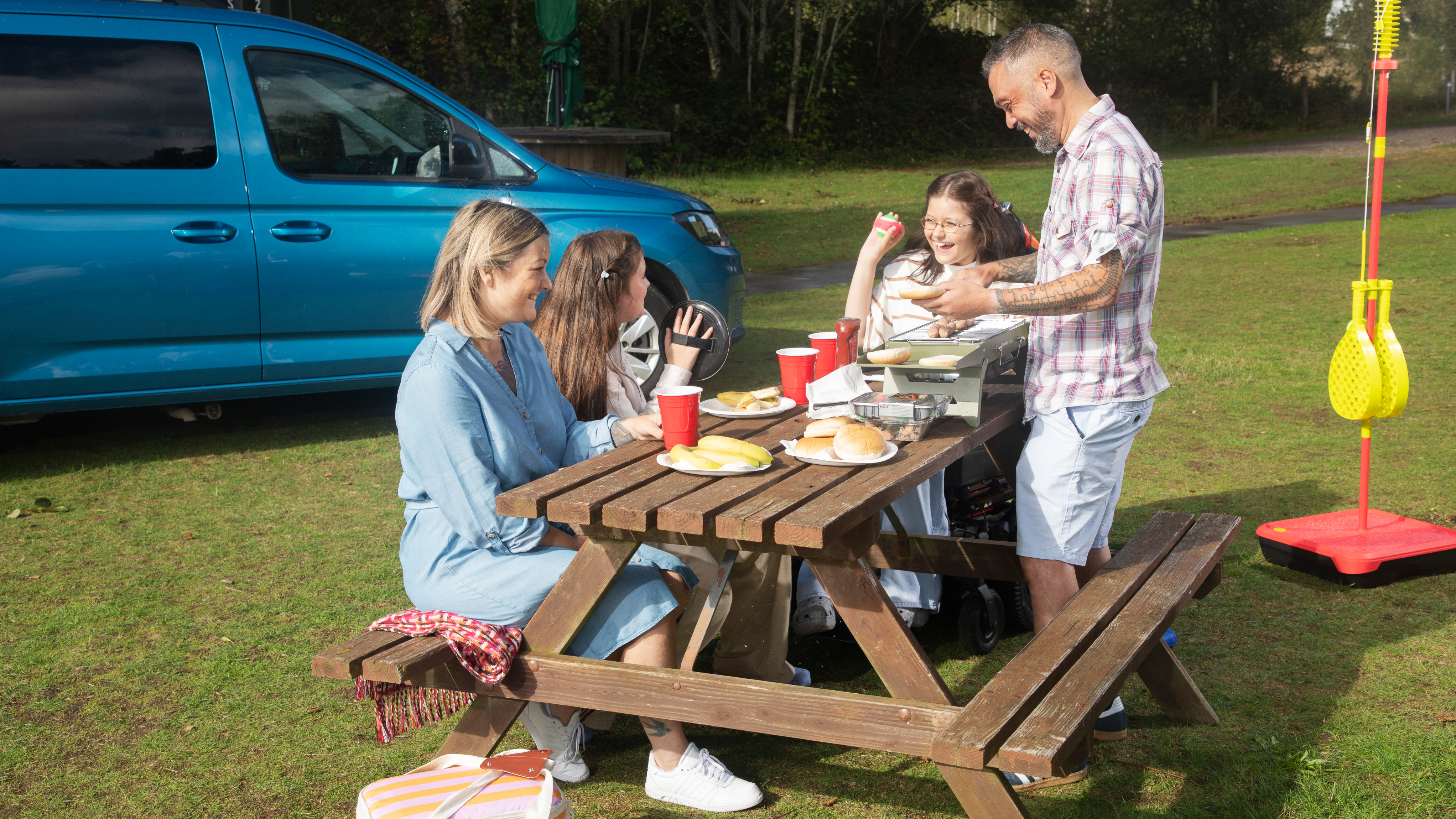 A family gathers around a wooden picnic table next to a blue accessible vehicle. Two adults and two children share food and laugh together in a grassy outdoor setting with play equipment nearby.