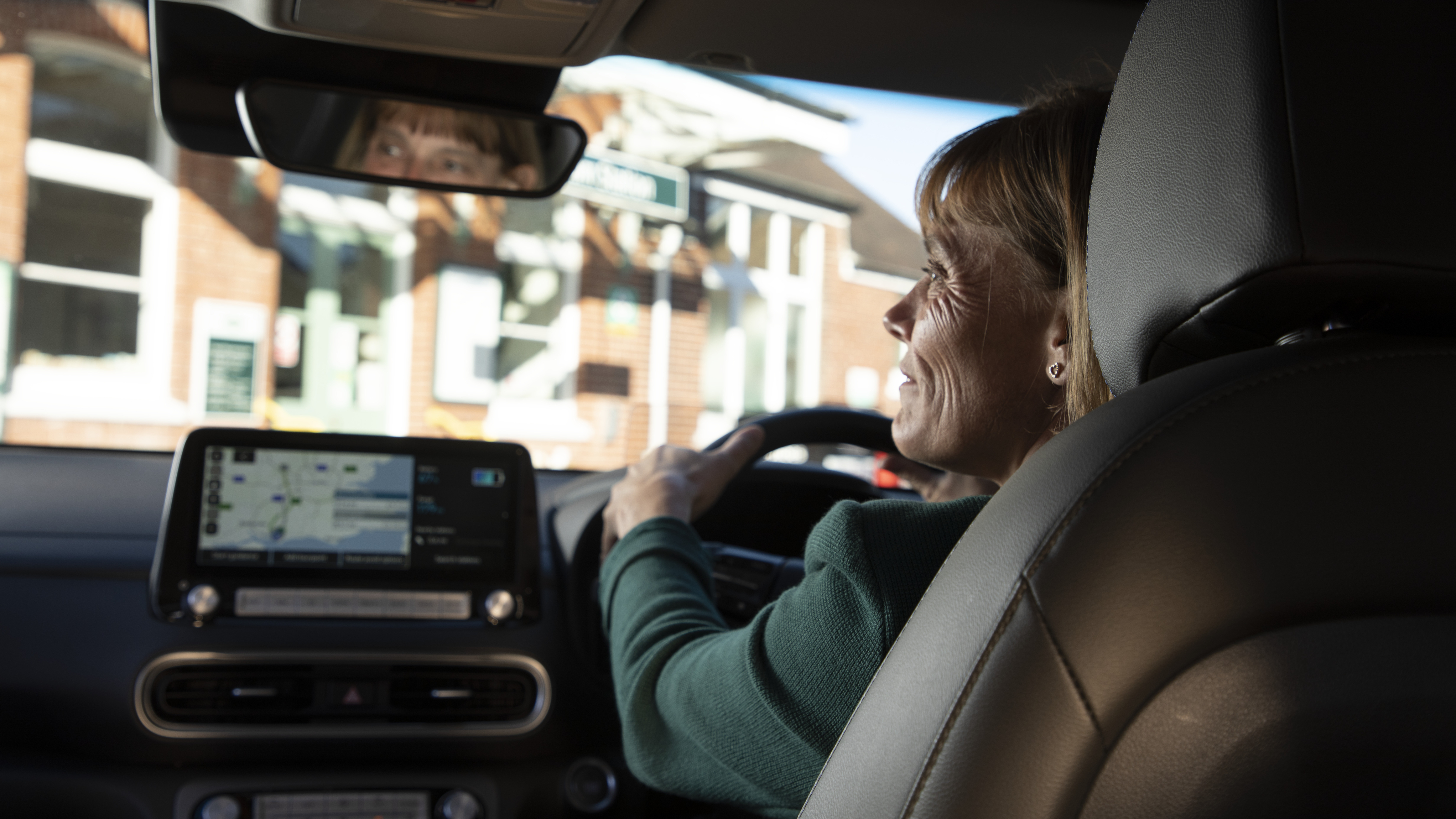 A woman in the driver’s seat of a car looks over her shoulder while reversing. The interior dashboard and navigation screen are visible, with sunlight streaming through the window and a brick building outside.