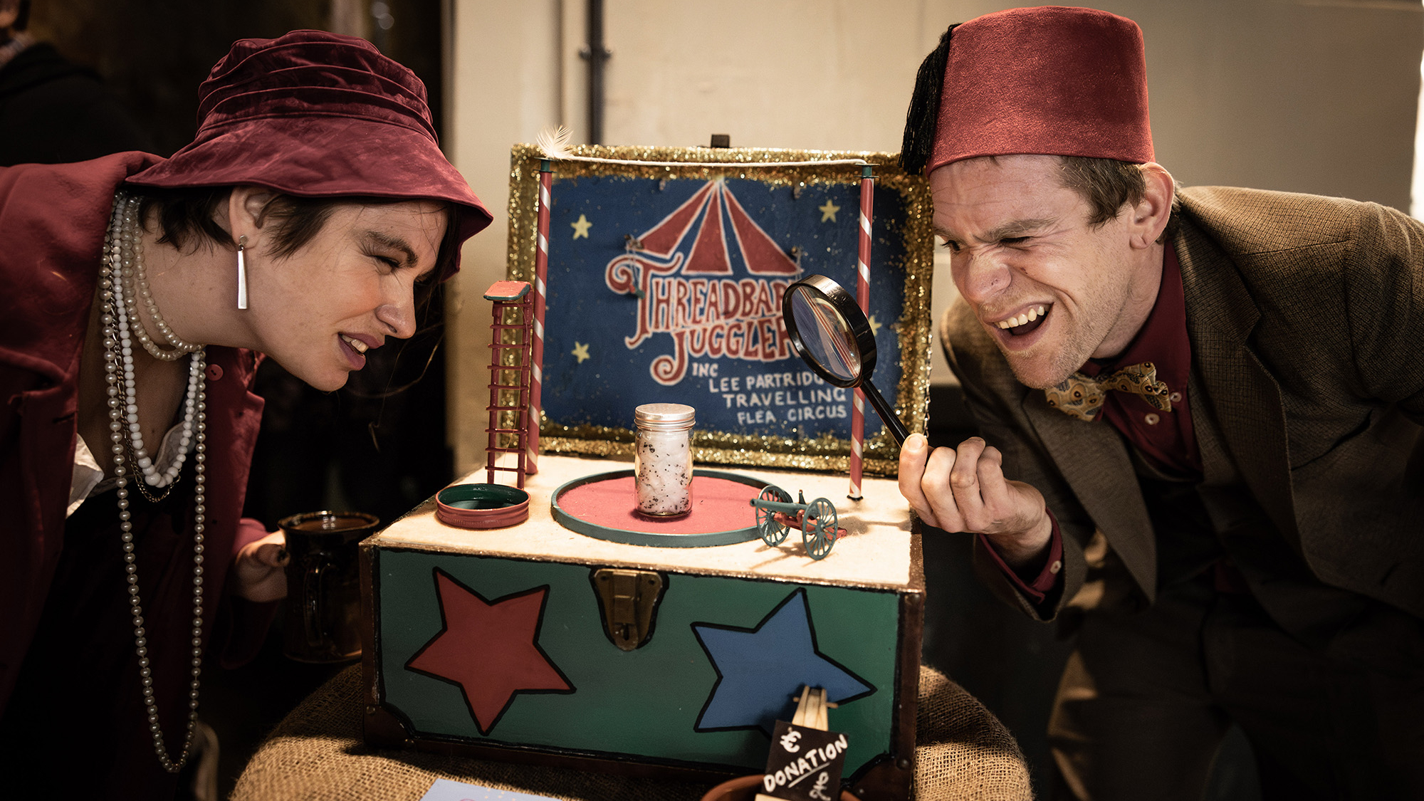 Two performers dressed in vintage costumes peer closely at a miniature circus display marked “Threadbare Jugglers,” using a magnifying glass to inspect tiny details.