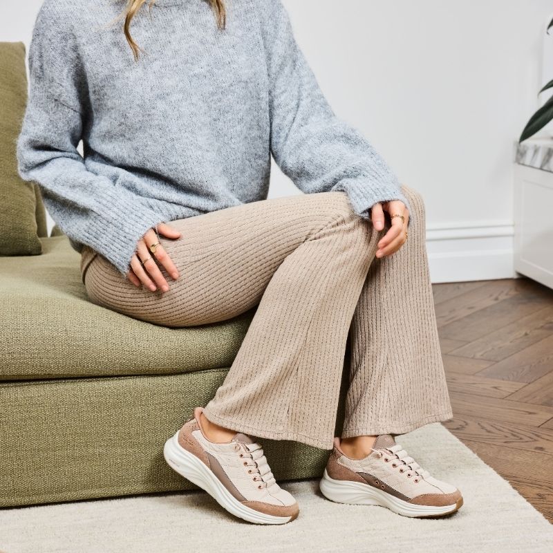 Person seated on a lounge wearing beige Skechers Cozy Fit sneakers with a grey knit jumper and ribbed pants.