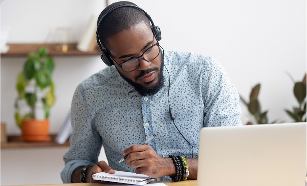 A focused, bearded man wearing glasses and a headset writes in a spiral notebook while looking at a laptop screen, likely participating in an online meeting or study session.