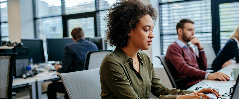 A focused woman with curly hair is working on a laptop in an open-plan office, with other colleagues visible working at their desks in the background.