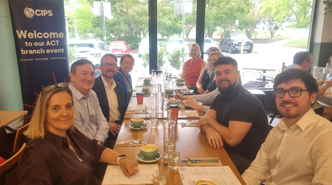 A group of people sitting around a long wooden café table with coffee cups, next to a banner that reads "Welcome to our ACT branch event."