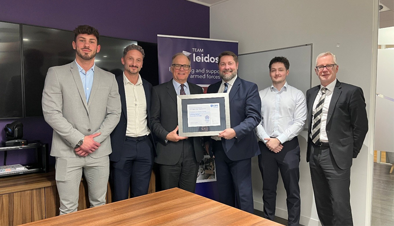 A group of six men in professional business attire stand in an office for a certificate presentation. Two men in the center hold a large, framed CIPS certificate. In the background, a purple banner for Team Leidos is visible, featuring the text Equipping and supporting our armed forces.