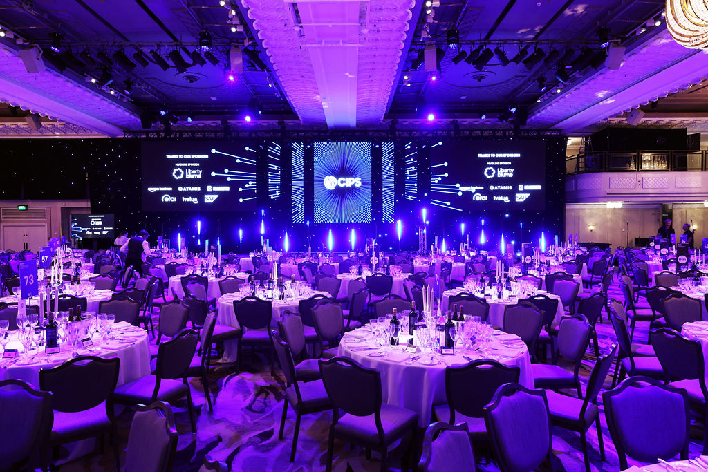 An expansive, grand ballroom is prepared for a CIPS (Chartered Institute of Procurement & Supply) awards ceremony, bathed in vibrant purple and blue lighting.