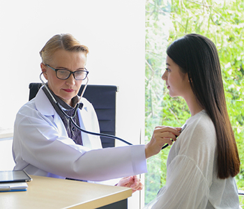 Starejša zdravnica v ordinaciji s stetoskopom posluša dihanje mlade pacientke.