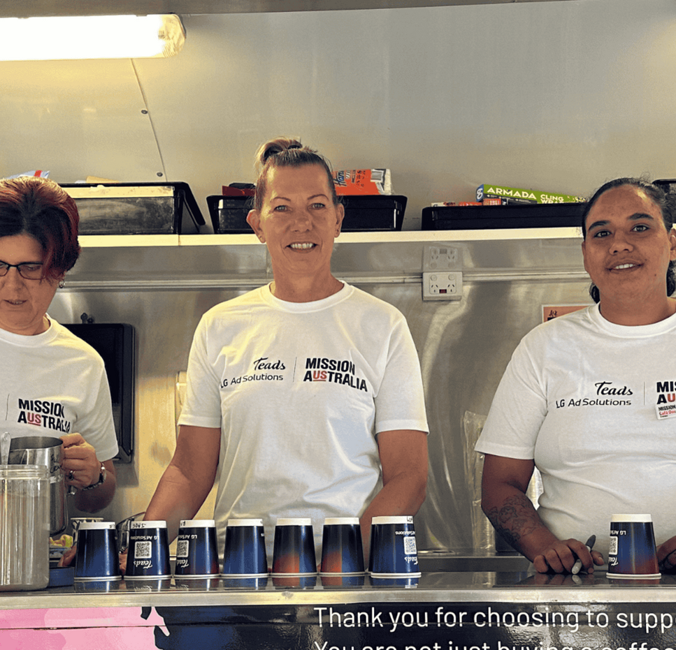 Three women wearing Mission Australia Café One shirts serve coffee from a colourful mobile café, supporting training and employment opportunities.