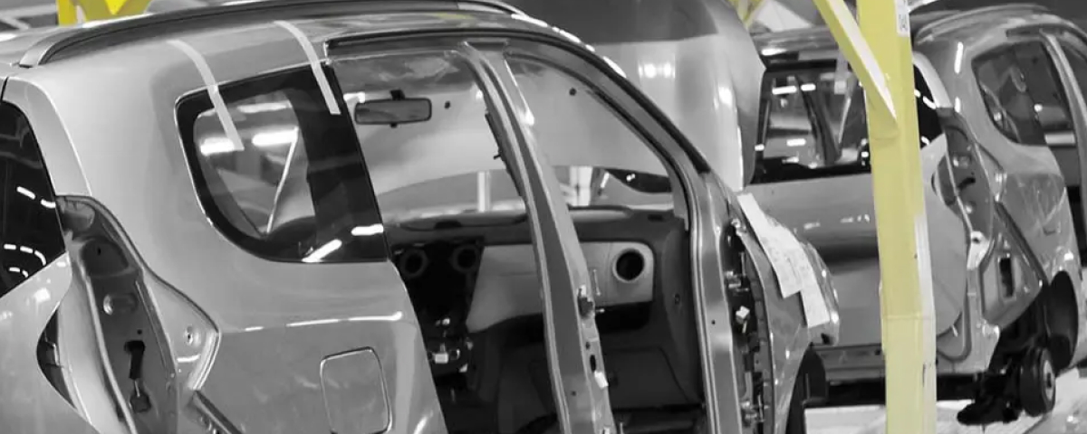 A black and white image of a car assembly line in a factory. Multiple car bodies are shown in various stages of assembly, mounted on a conveyor or transport system. The nearest car is shown from the side, with its doors open. The only color present is the bright yellow of the structural supports and railings of the factory equipment, which provides a visual contrast to the grayscale vehicles.