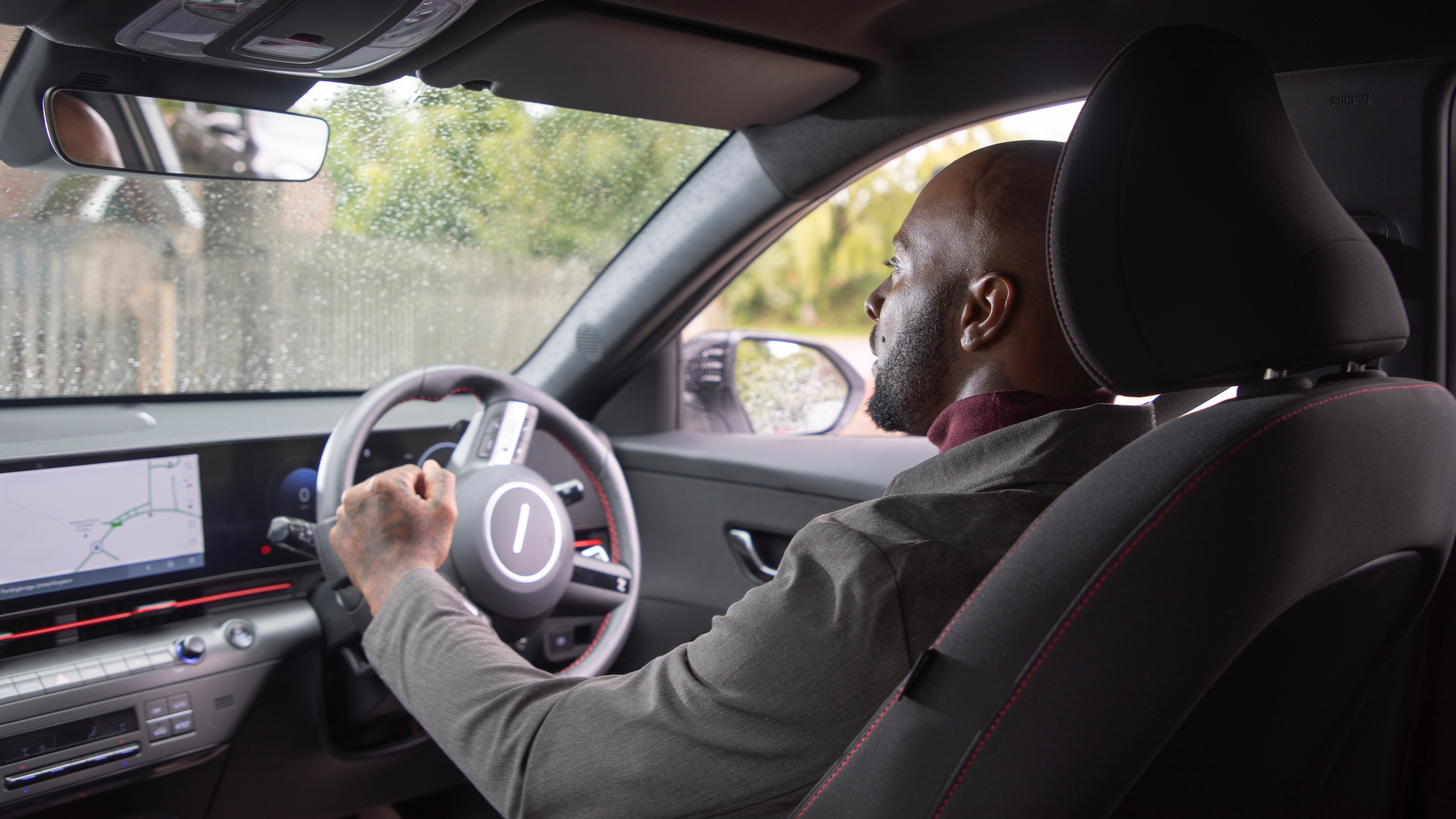 A man sits in the driver’s seat of a modern car, holding the steering wheel and looking ahead. The interior shows a digital dashboard and infotainment screen, with raindrops visible on the window outside.