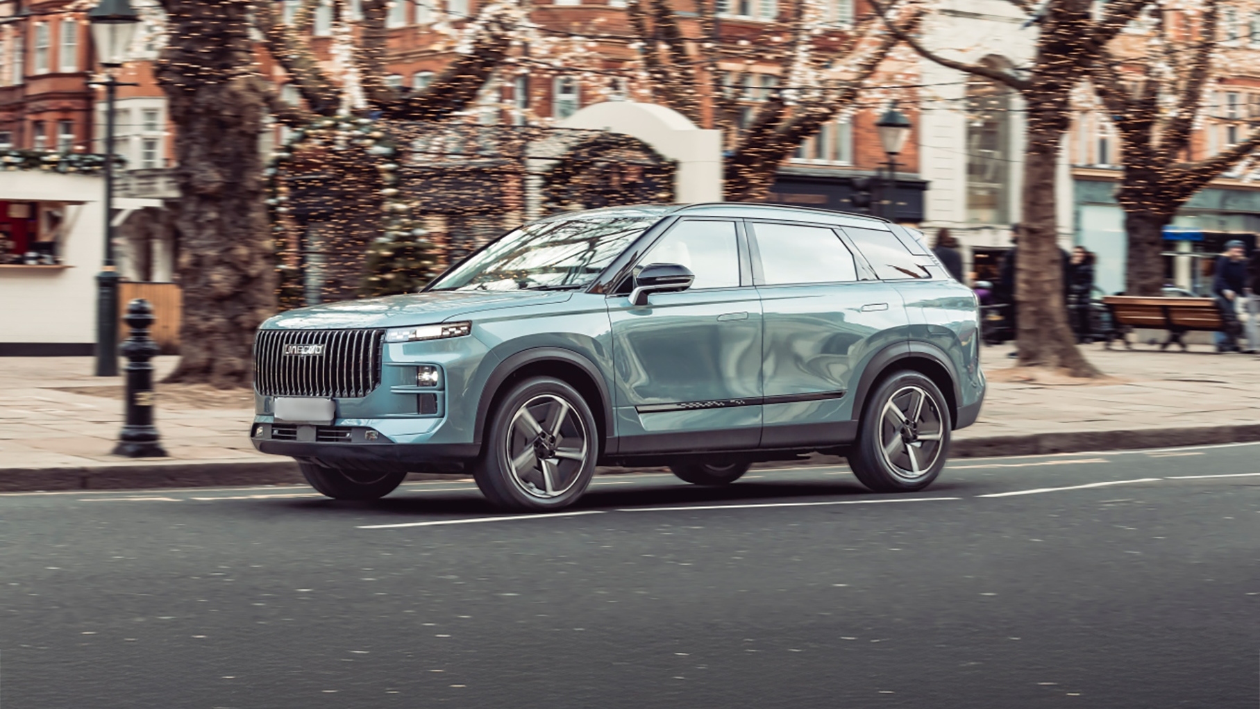 Light blue Jaecoo 7 SUV driving through a city street decorated with lights, shown from the side front angle.