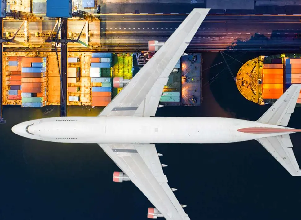 Aerial view of a white airplane flying over a colorful cargo port with containers and ships.