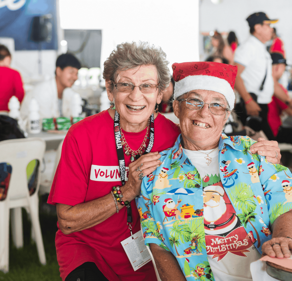 Two smiling individuals pose at a festive event. One wears a red "VOLUNTEER" shirt, and the other dons a Santa-themed shirt and hat.