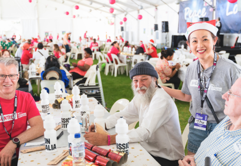 Volunteers and guests sit at a table with Mission Australia water bottles during a Christmas lunch event under a decorated tent.