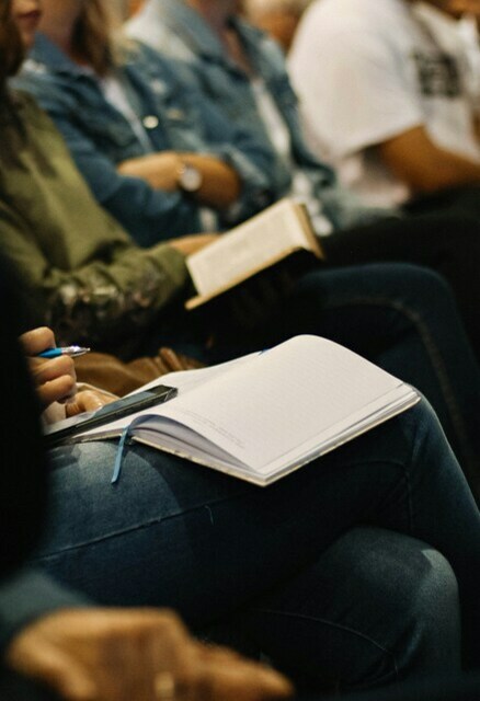 A group of people seated closely together, some holding open notebooks and pens, suggesting they are taking notes during a lecture or seminar. The focus is on their laps and hands, with faces out of focus.