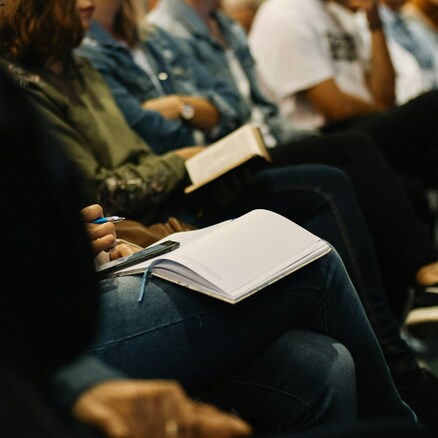 A group of people seated closely together, some holding open notebooks and pens, suggesting they are taking notes during a lecture or seminar. The focus is on their laps and hands, with faces out of focus.
