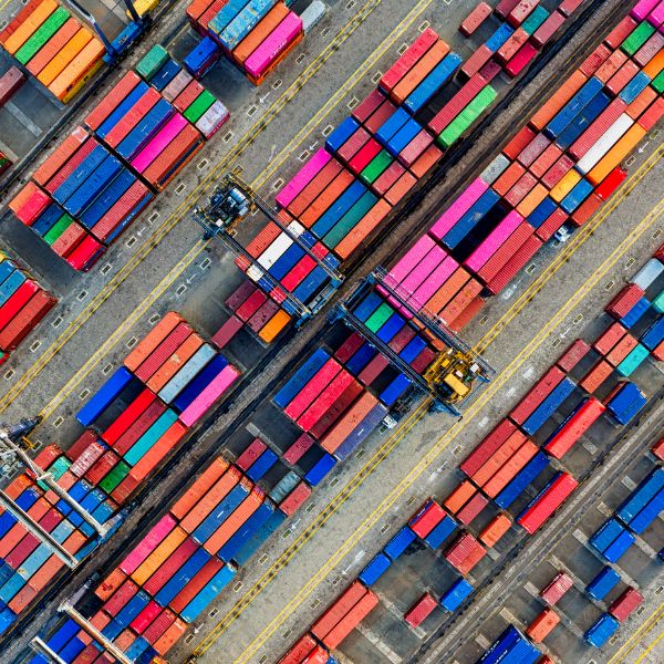 High-angle aerial view of a shipping port featuring rows of colourful red, blue, and orange shipping containers organized in a geometric pattern with a yellow gantry crane.