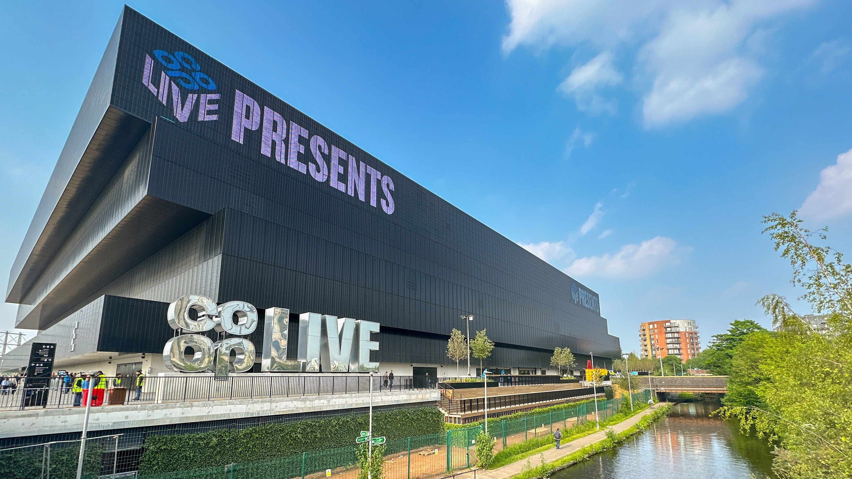 Exterior of Co-op Live arena in Manchester with large “Co-op Live Presents” signage on the building, set beside a canal and walkway with people nearby under a clear blue sky.