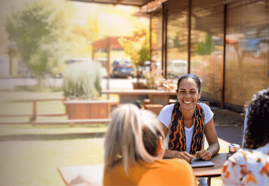 A woman smiles while sitting at an outdoor table with three other people, enjoying a sunny day. She has a notebook in front of her and appears to be engaged in a friendly conversation.