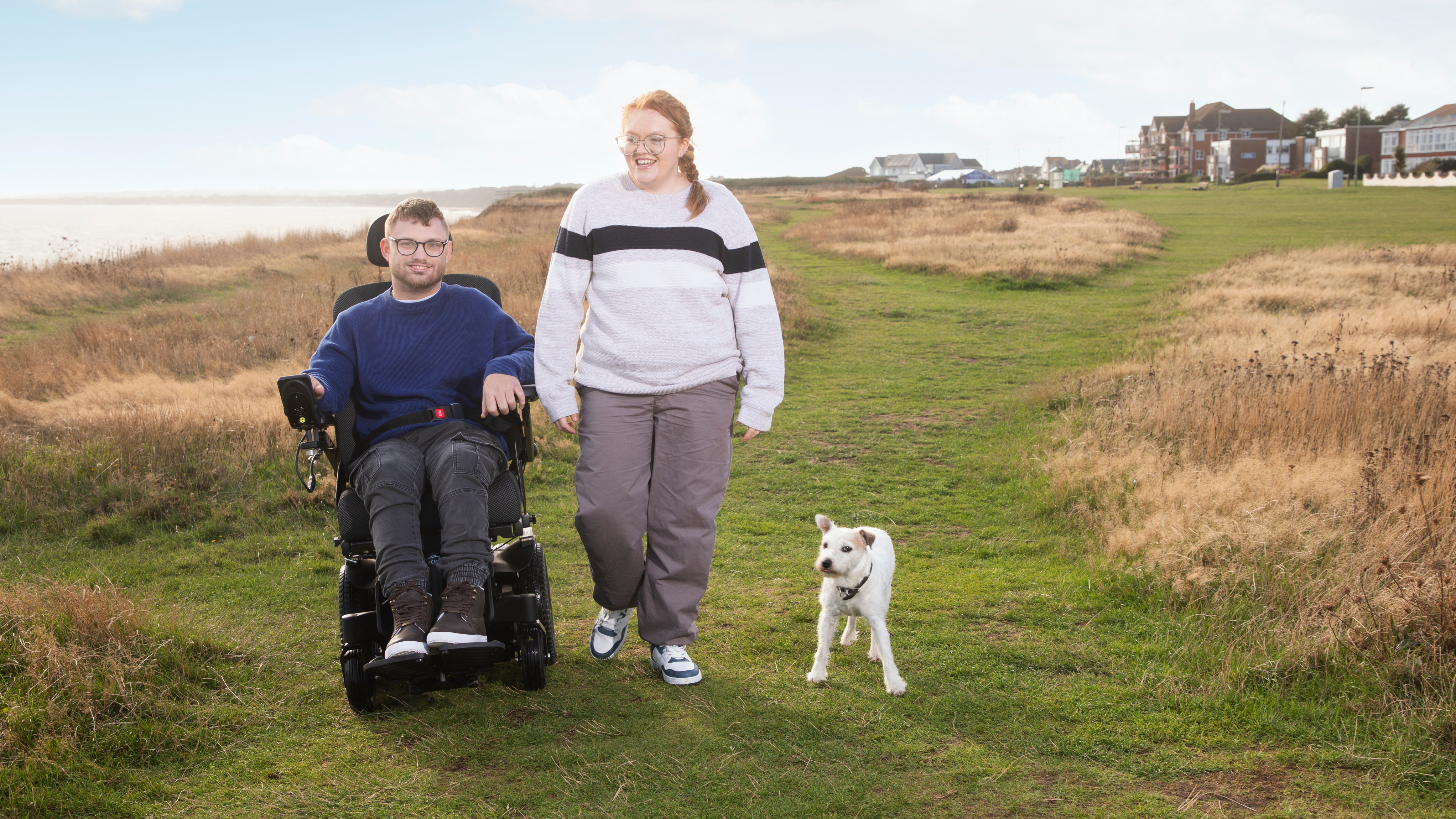 A man using a powered wheelchair walks alongside a woman on a grassy coastal path, with a small white dog walking ahead and houses visible in the distance.