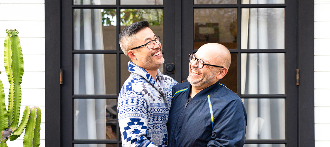 An Asian man and a white man stand hugging and laughing together in front of their home
