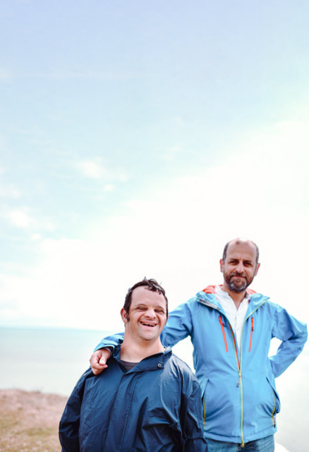 Two men standing on a cliff, smiling at the camera. One of the men appears to have a disability. 