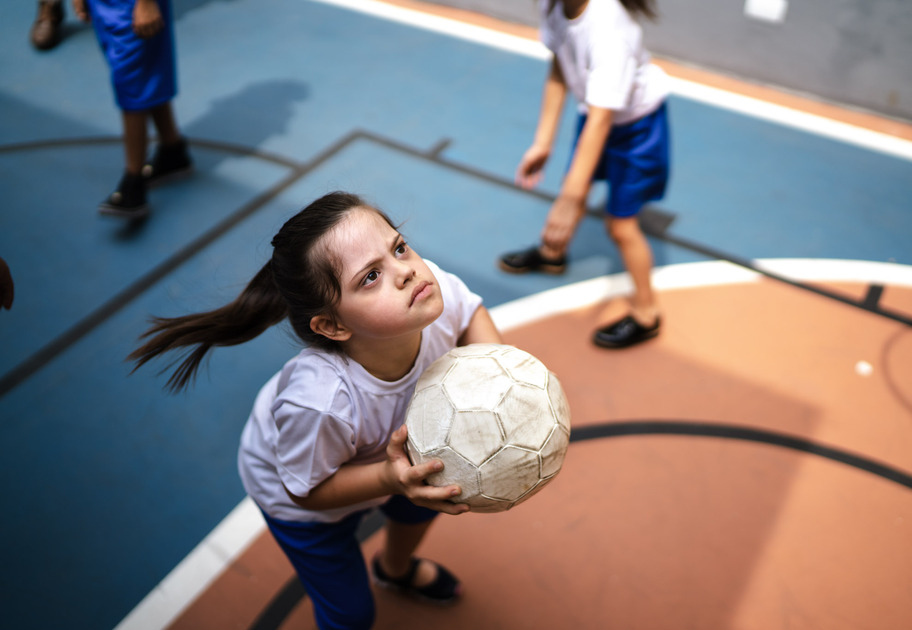 A young woman, who appears to have a disability, playing netball outdoors with her team. She looks determined as she takes a shot at the goal.