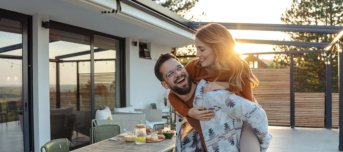A white man holds a white woman on his back as they laugh together on their porch