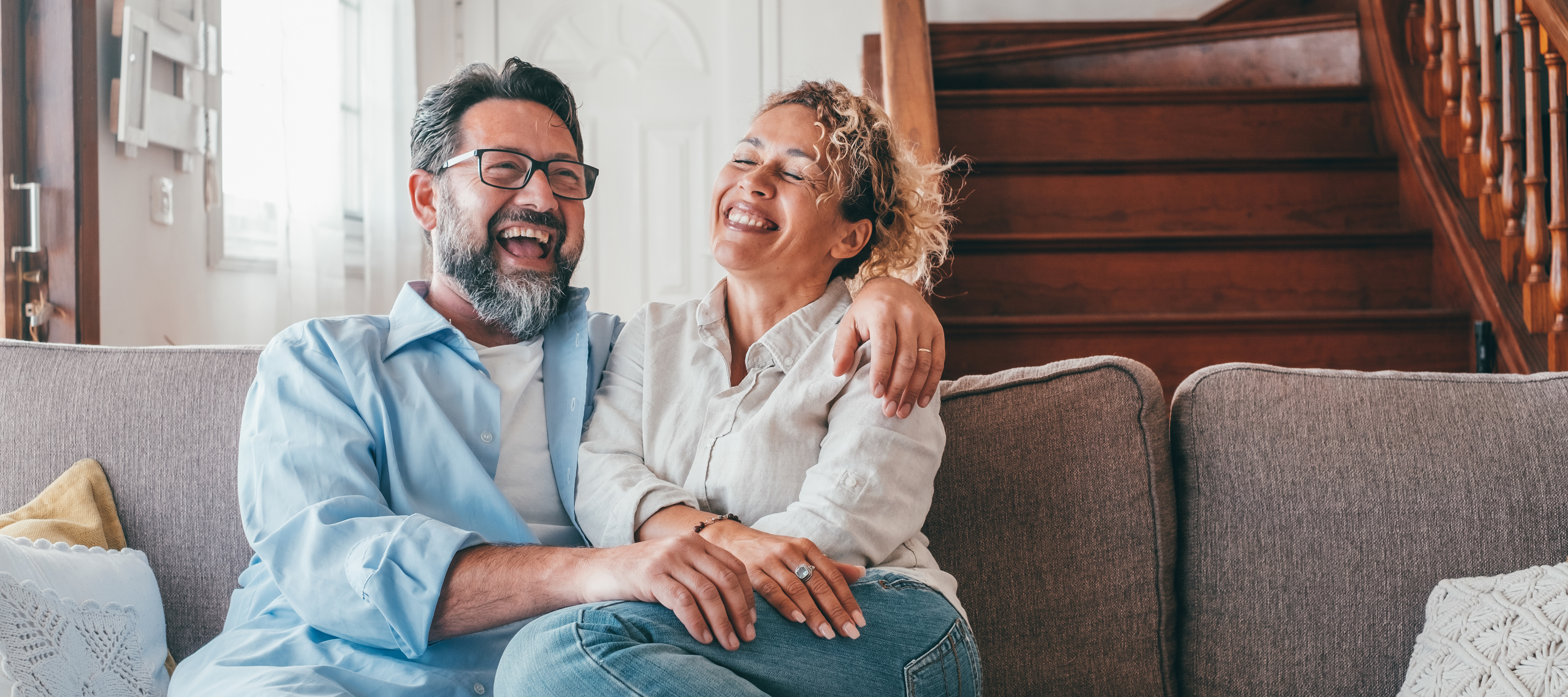 A man and woman sit laughing together on a couch in a nice house
