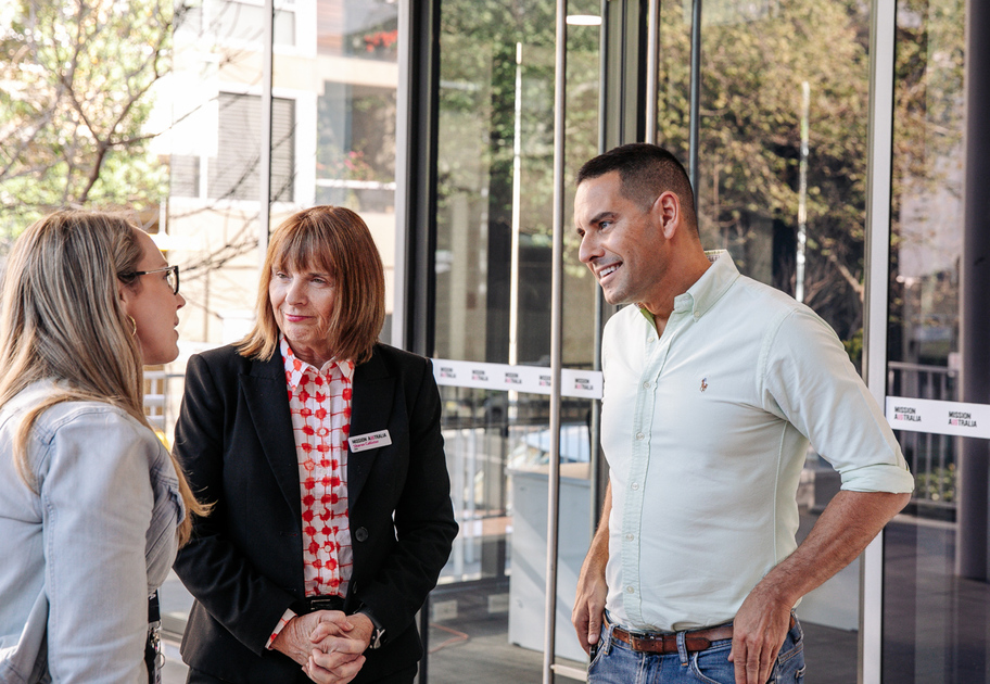 Three people are standing and talking in a modern building lobby with large glass windows. Two women, one in a suit and the other in a light jacket, face a man in a light green shirt who is smiling.