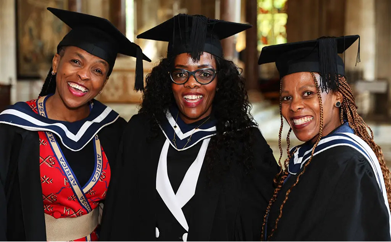 Three women in graduation caps and gowns are smiling and posing together for a photo indoors, likely in a large hall or church, with stained glass visible in the background. The woman on the left is wearing a red and black patterned garment underneath her gown.